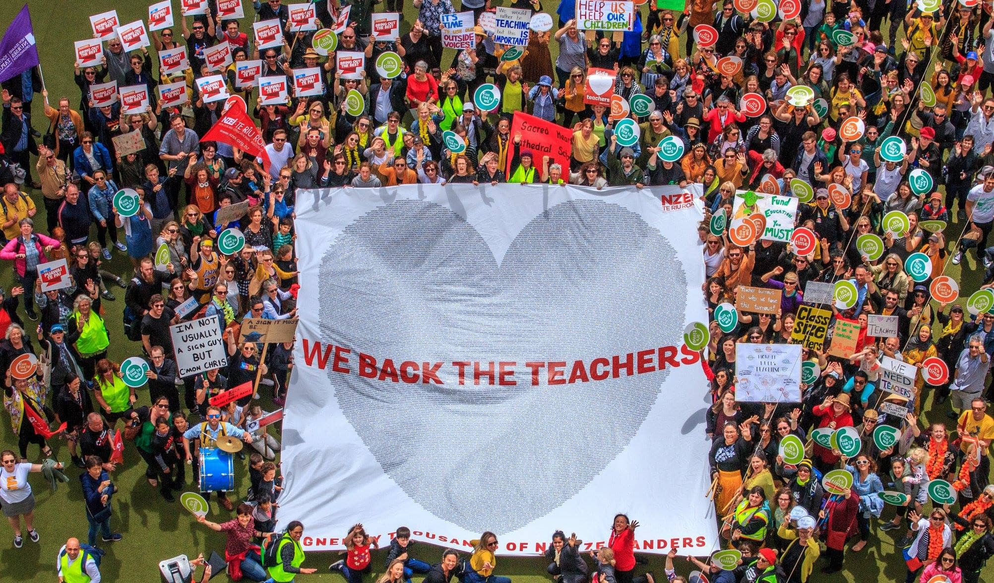 Huge and colorful crowd of union and teacher supporters in a field with a banner reading “We back the teachers”
